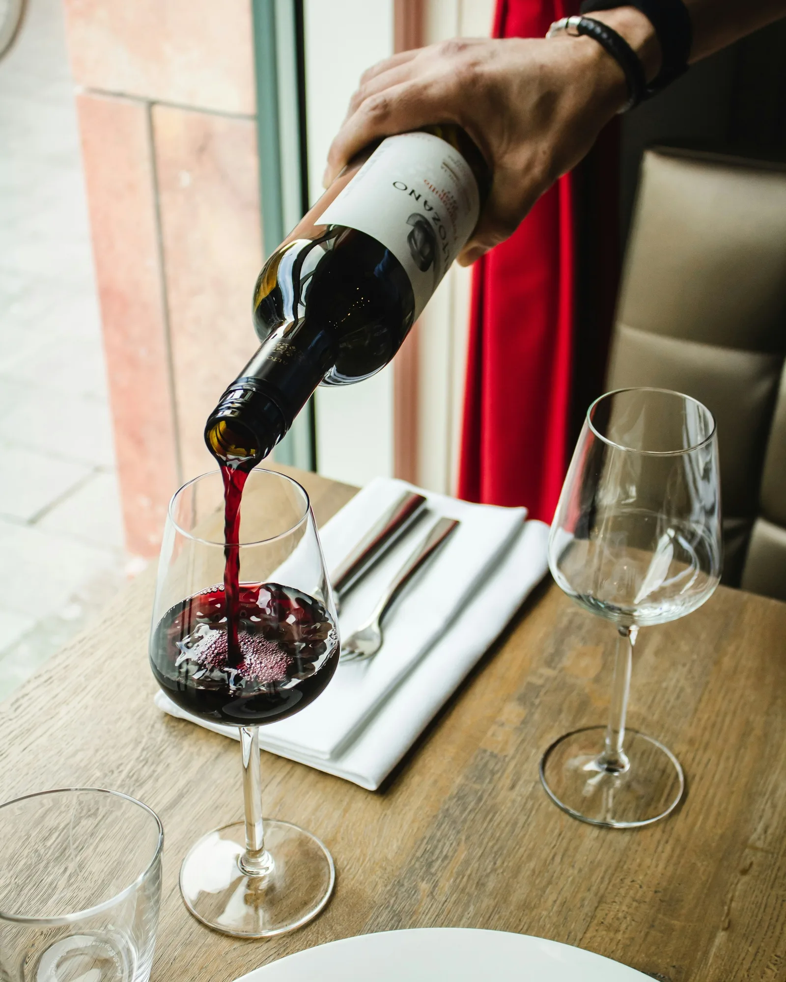 Uma pessoa serve vinho tinto num copo de vinho, sobre uma mesa de madeira onde estão dispostos copos vazios, um guardanapo branco e um prato com comida, junto a uma janela com luz natural