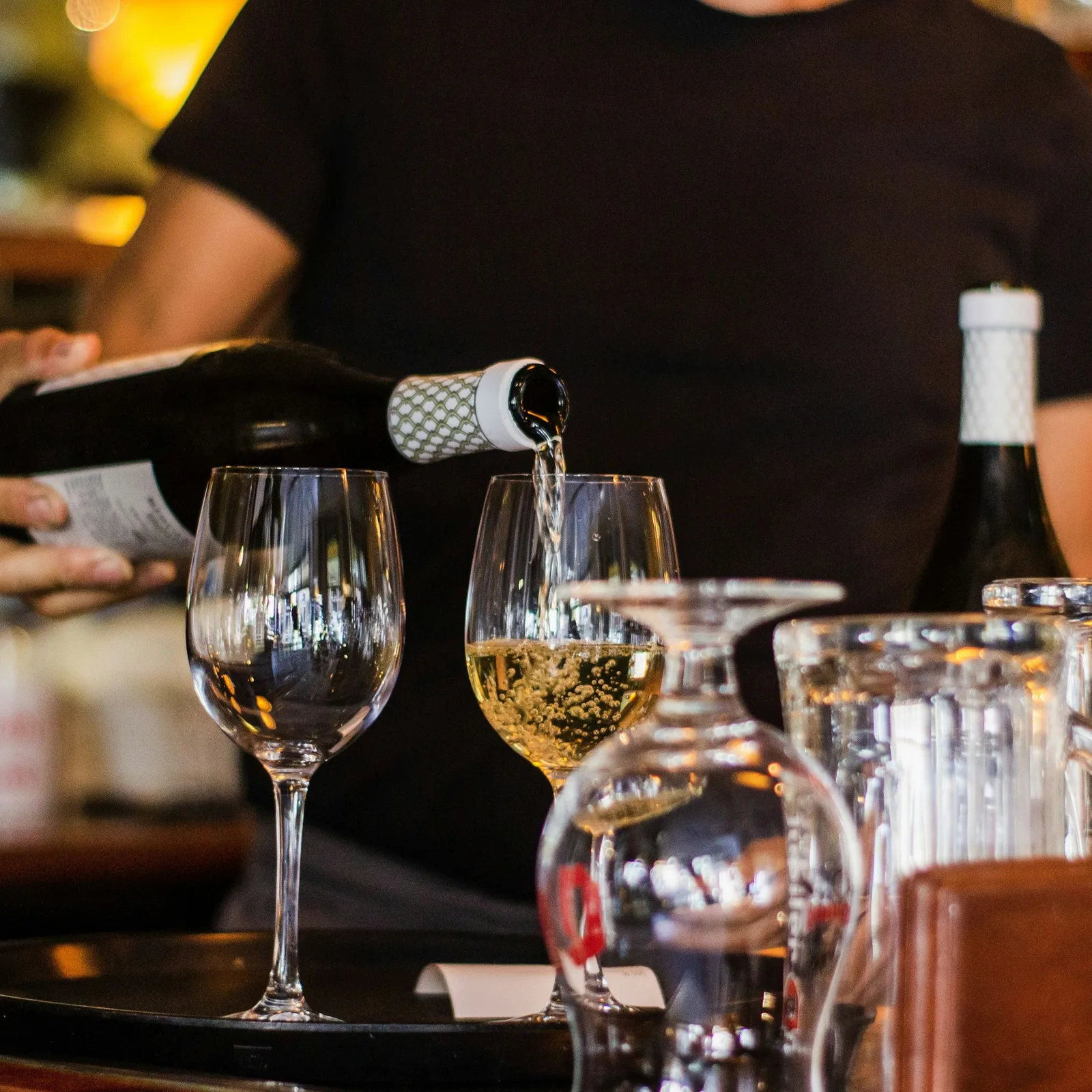 Uma pessoa a servir uma garrafa de vinho espumante num copo num bar ou restaurante, com vários outros copos e garrafas no balcão